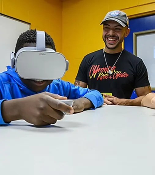 Dr. Danny Pimentel: laughing beside a young visitor trying a VR headset at a classroom table