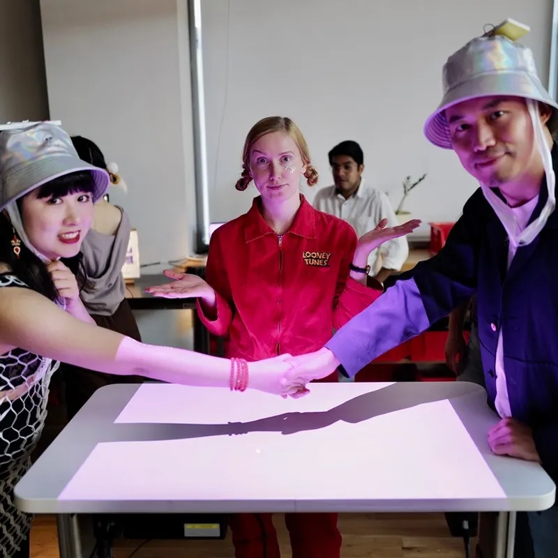 Kami Karras: a Spatial Design Club session: Kami in a red jacket between two friends in silver bucket hats shaking hands over a glowing table