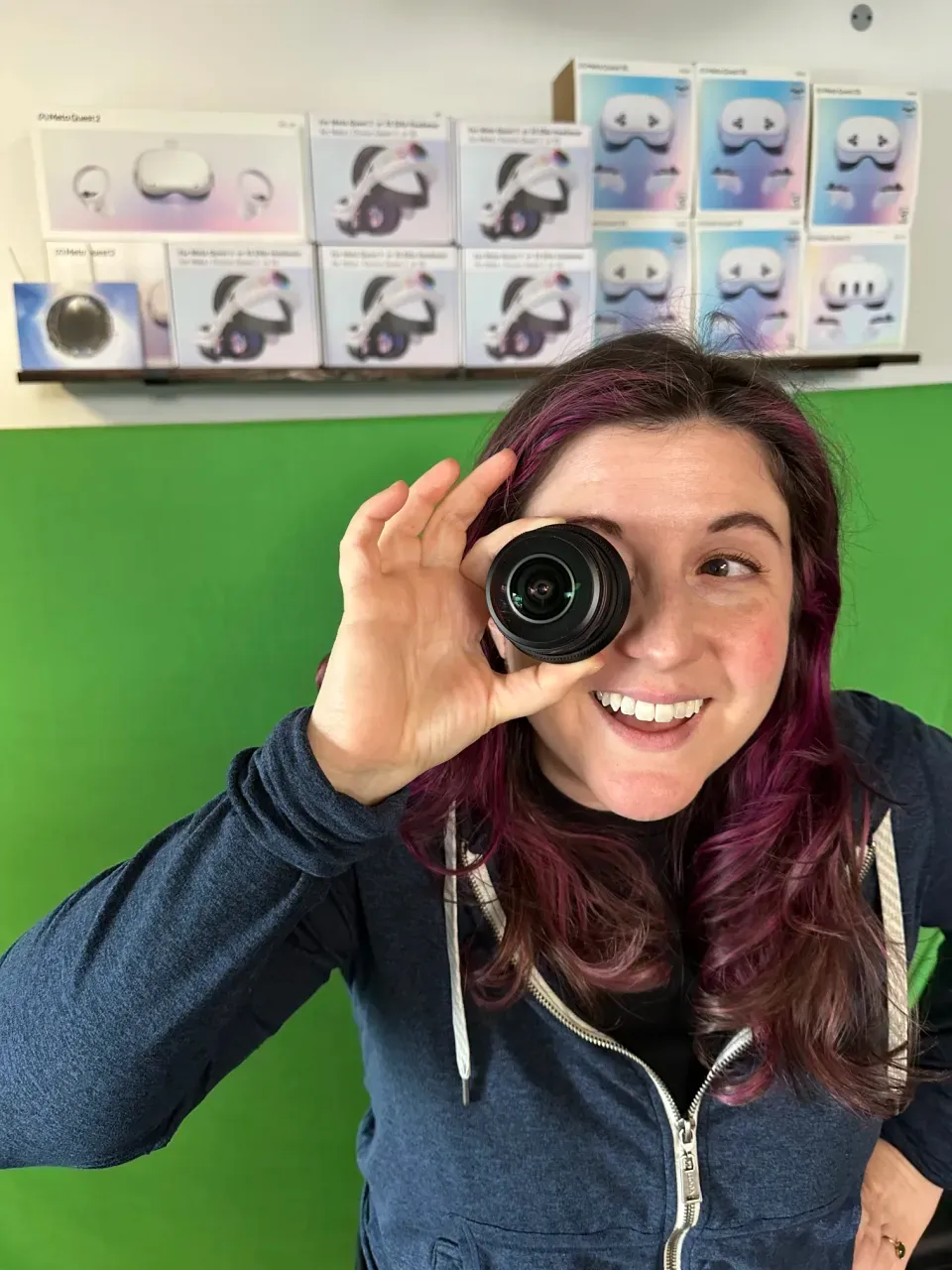 Rachel Hengchua: holding a camera lens up to one eye and smiling, in front of a shelf of VR headset boxes