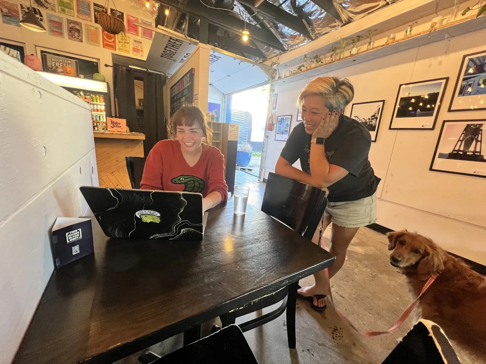 Heather Dunaway Smith and Estella Tse laughing over a laptop at a table inside Daydreamer Coffee & Public House, with Estella’s dog Bryan beside them