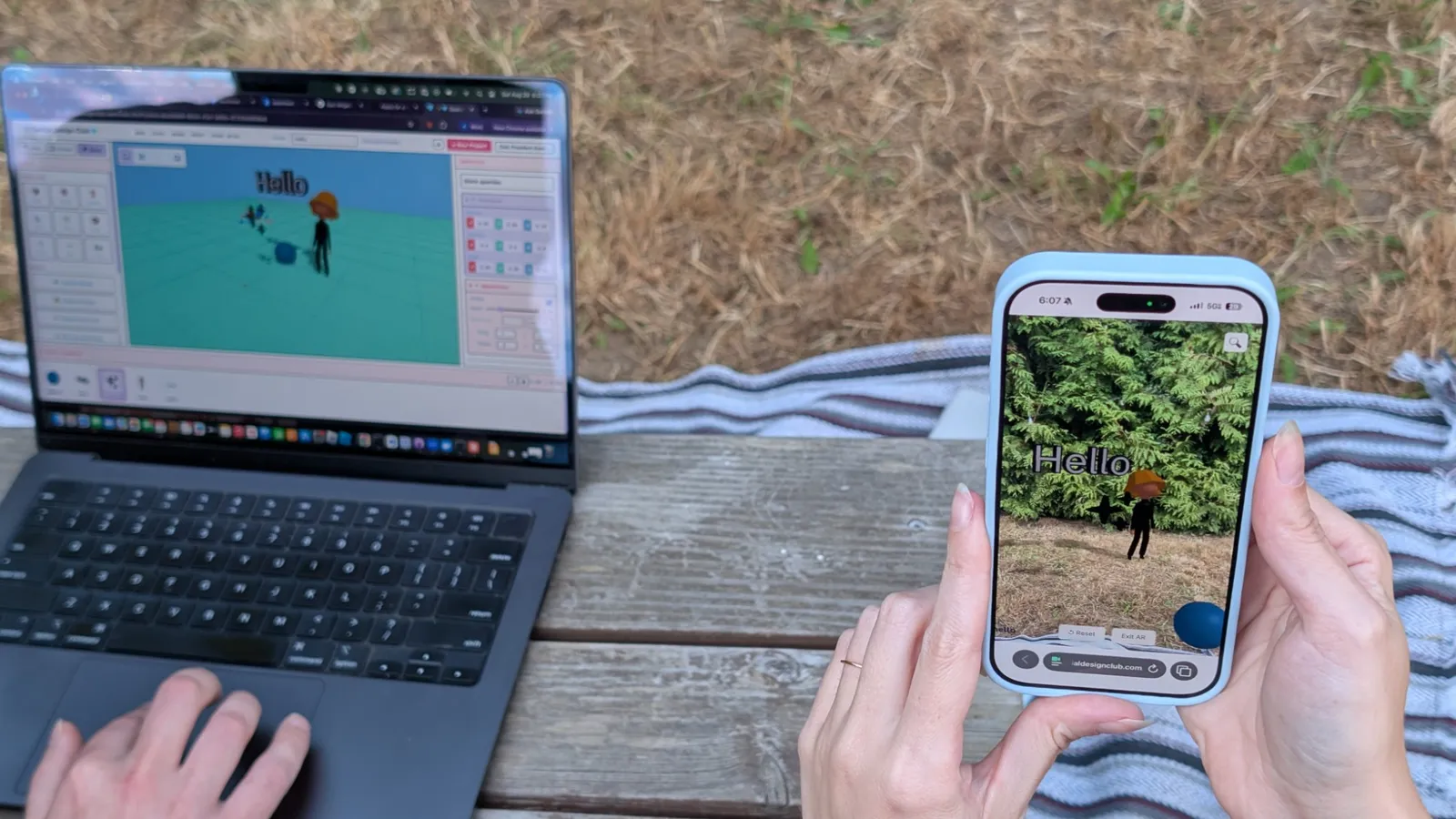 A laptop at a picnic table shows a scene being built in Spatial Design Club while a phone held up beside it shows the same scene placed in the grass in AR